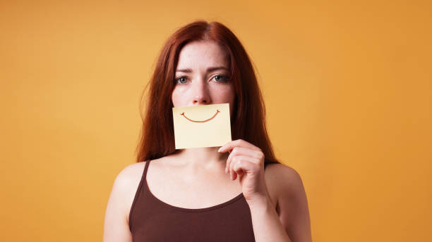 Woman hiding her real smile behind a paper with a drawn smile — emotional toll of dental issues